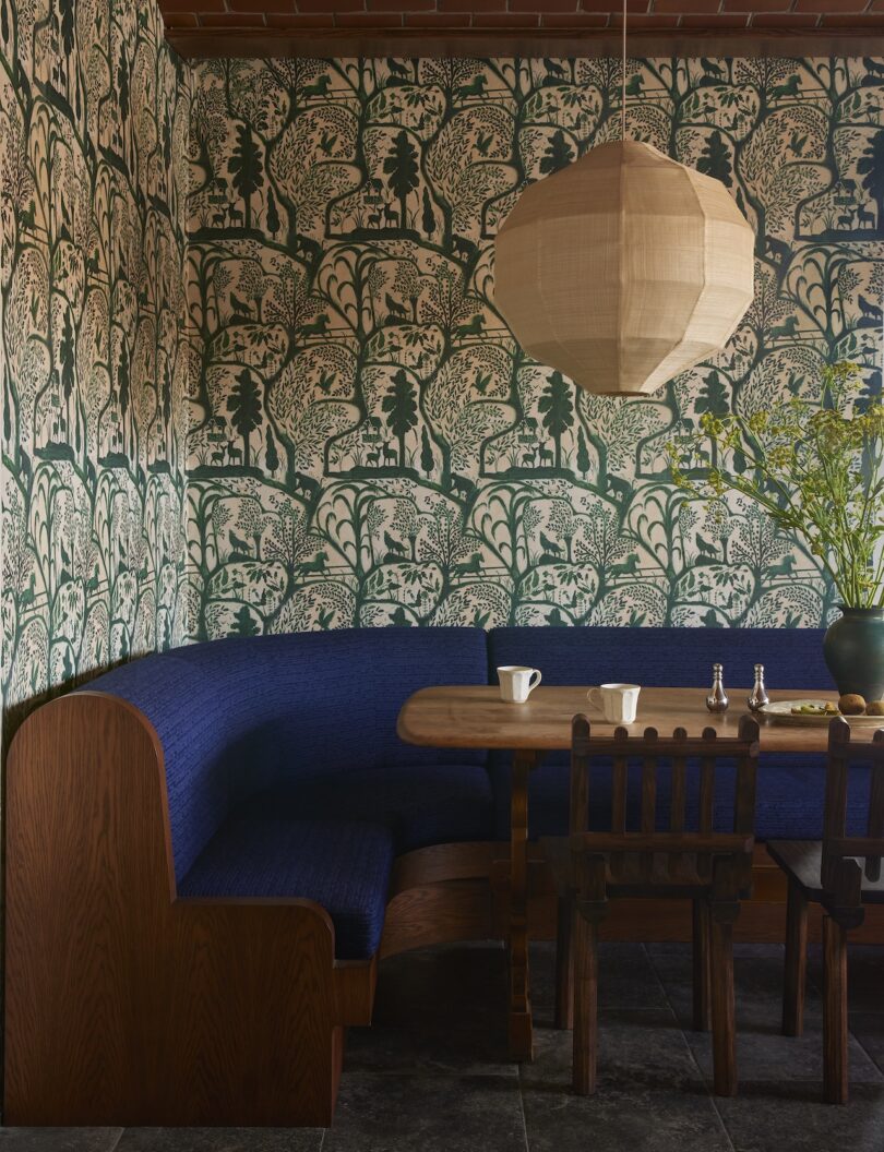 A dining nook with a curved blue upholstered bench, a wooden table and chairs, green patterned wallpaper, a paper lantern and a vase of branches.