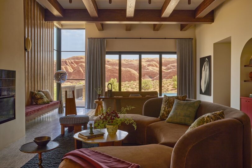 A living room with brown curved sofas, a round wooden coffee table, large windows overlooking the desert cliffs and exposed wooden beams on the ceiling.