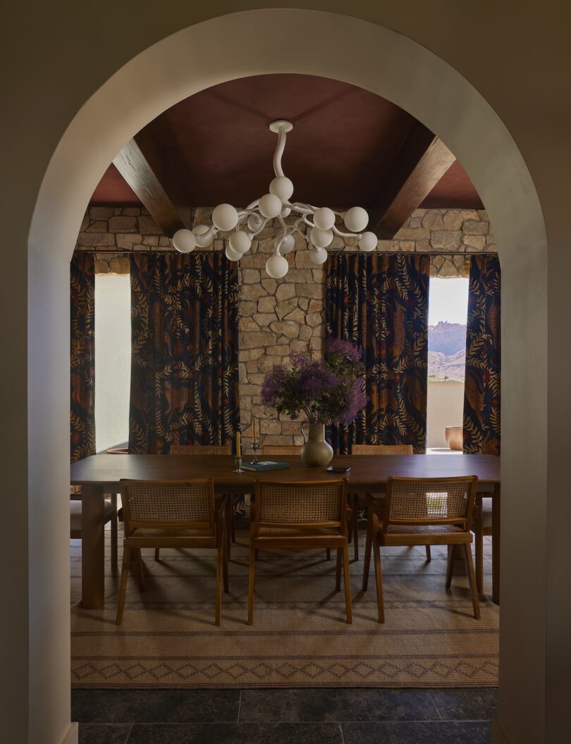 A dining room with a wooden table, six chairs, a modern white chandelier, stone walls, patterned curtains and a flower vase, viewed through an arched doorway.