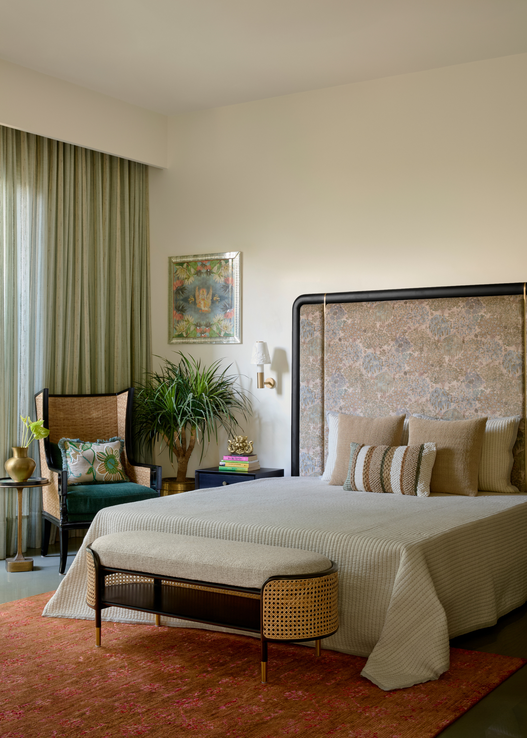 A cream bedroom with an oversized headboard, a set of beige textured pillows, a rattan-style bench and a potted plant in a gold planter next to a rattan chair
