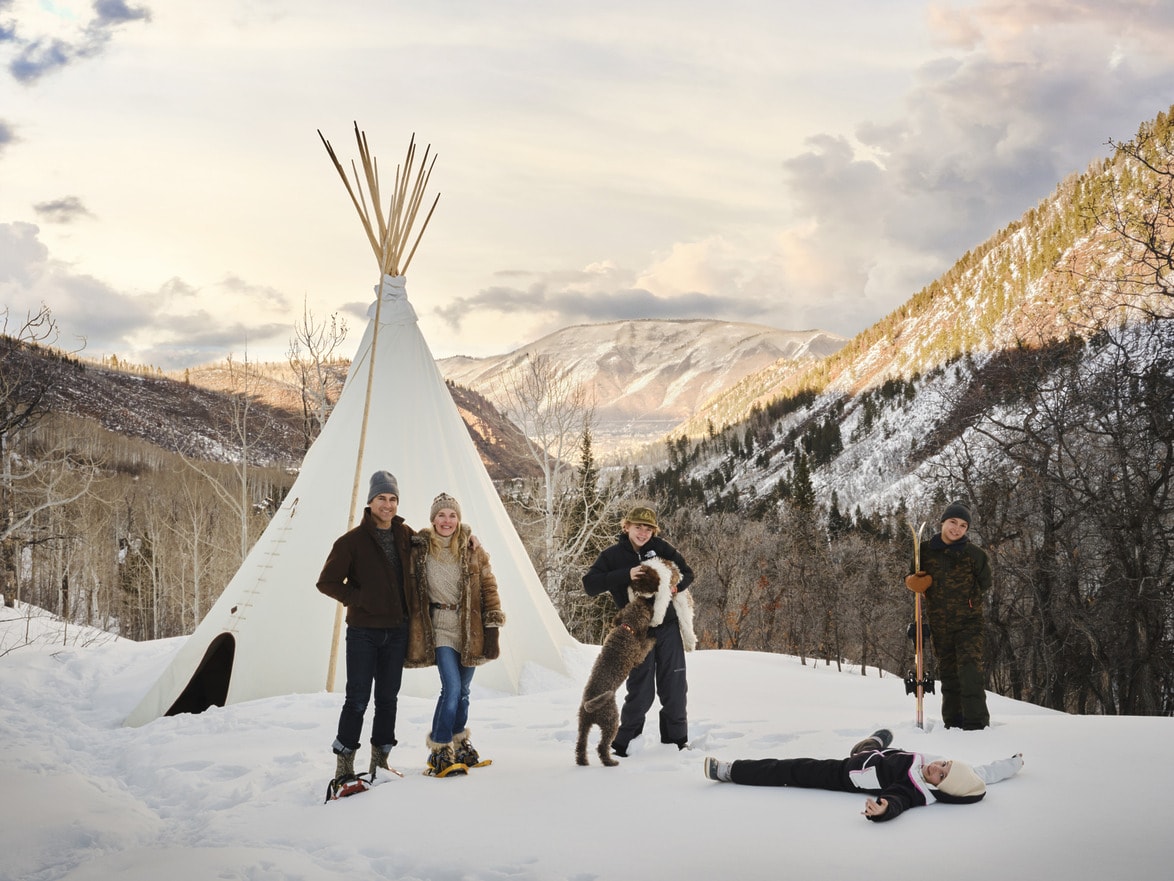 Family in winter equipment with dog in front of teepee in snowy mountain landscape at sunset.