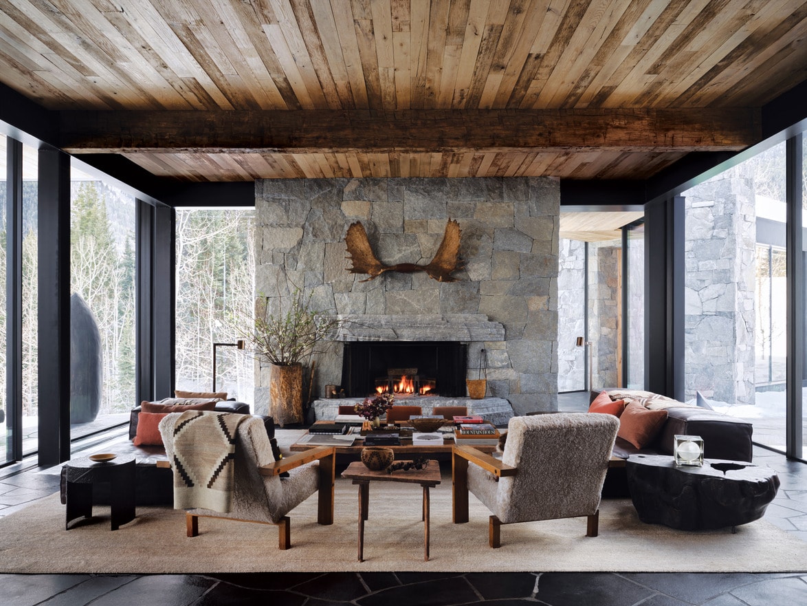 Cozy living room with stone fireplace, wooden ceiling and large windows overlooking the snowy forest landscape.
