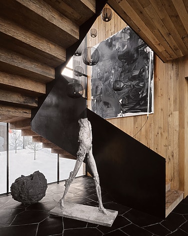 Modern art sculpture of a faceless human figure under a staircase surrounded by abstract paintings and wooden walls.