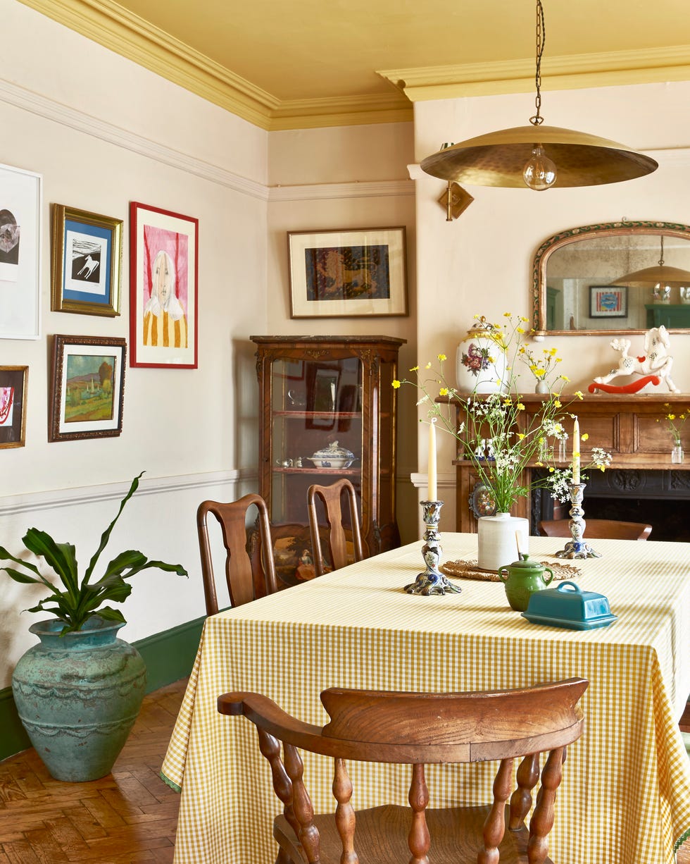 a dining area with a yellow gingham dining table and wooden chairs, a wooden cupboard and light yellow walls with wall art on them