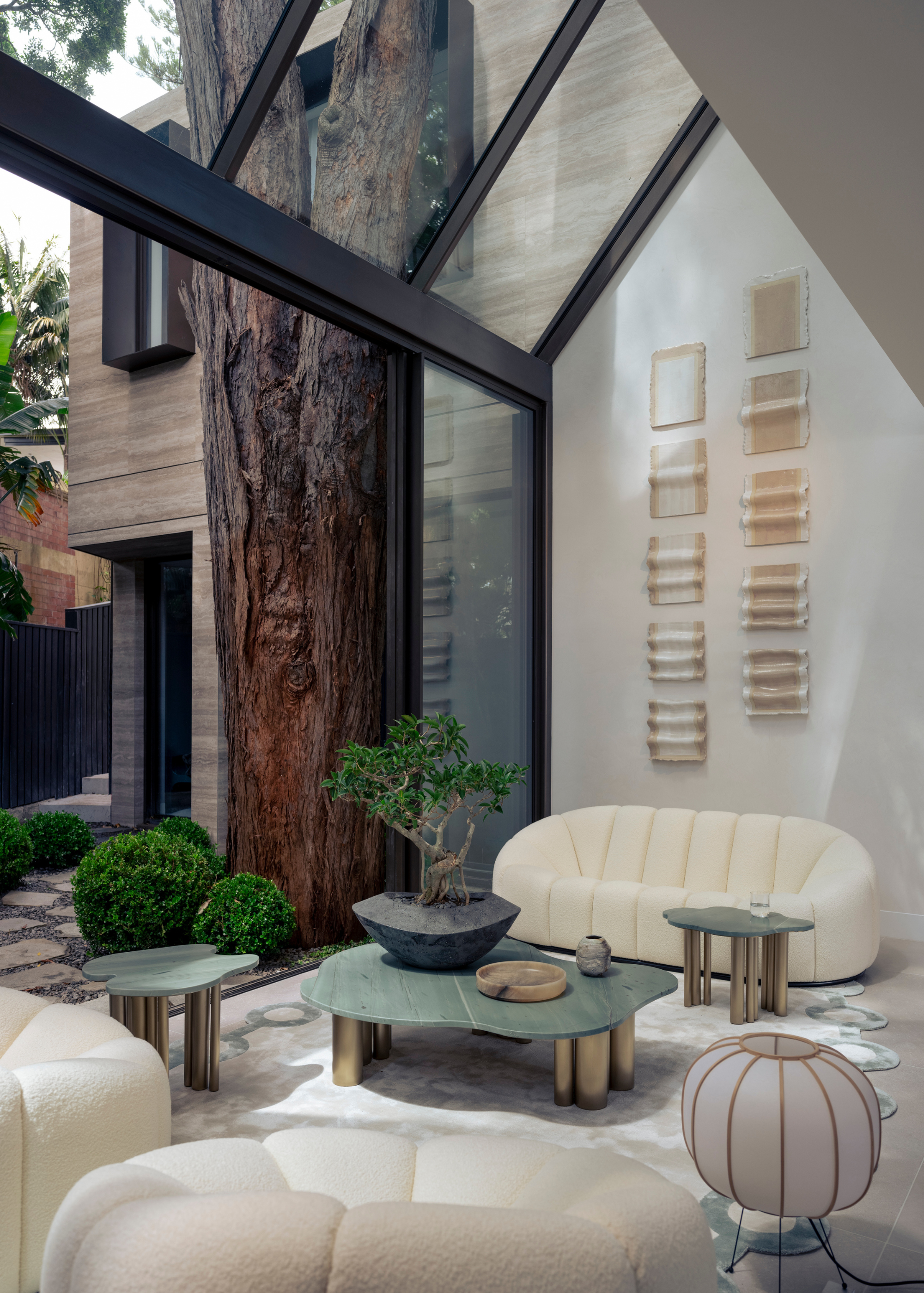 A white living room with vaulted window ceilings, a bonsai in a black planter on a green coffee table next to a white couch and sofa seats, a rice paper floor lamp and textured wall hangings
