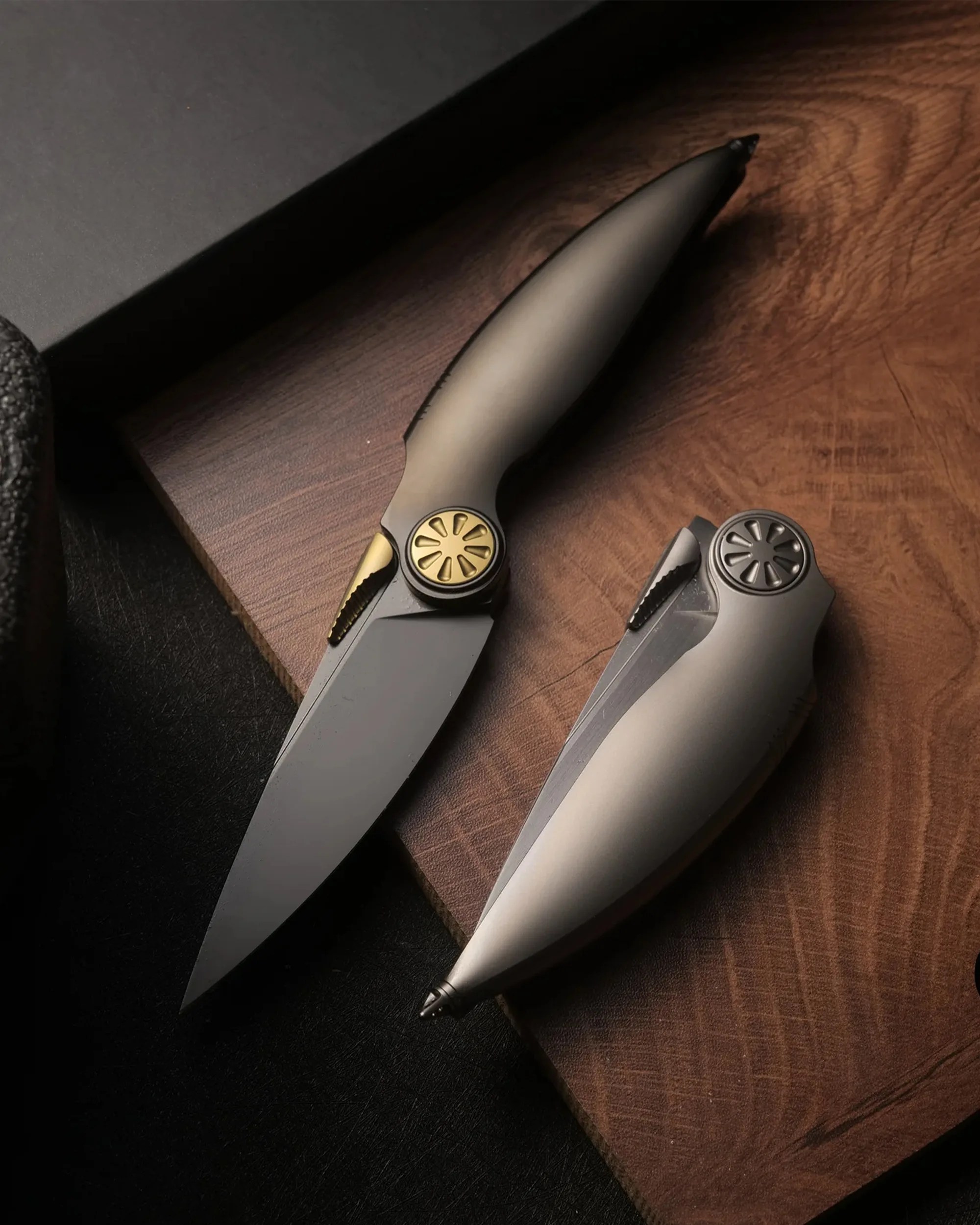 Two elegant folding knives with matte gray handles and gold and black accents on a wooden surface.