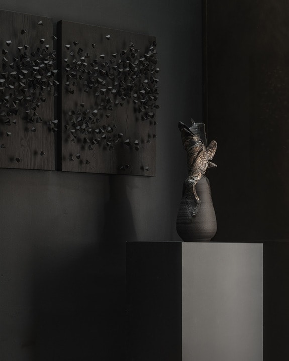 Dark modern art installation featuring sculptures on a pedestal and textured wall panels in a dimly lit room.