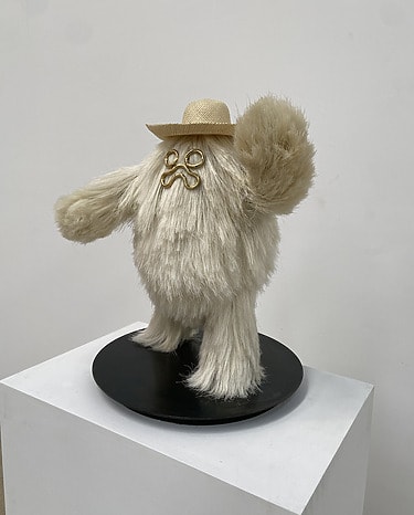 Sculpture of a furry creature wearing a hat and glasses on a black base against a white background.