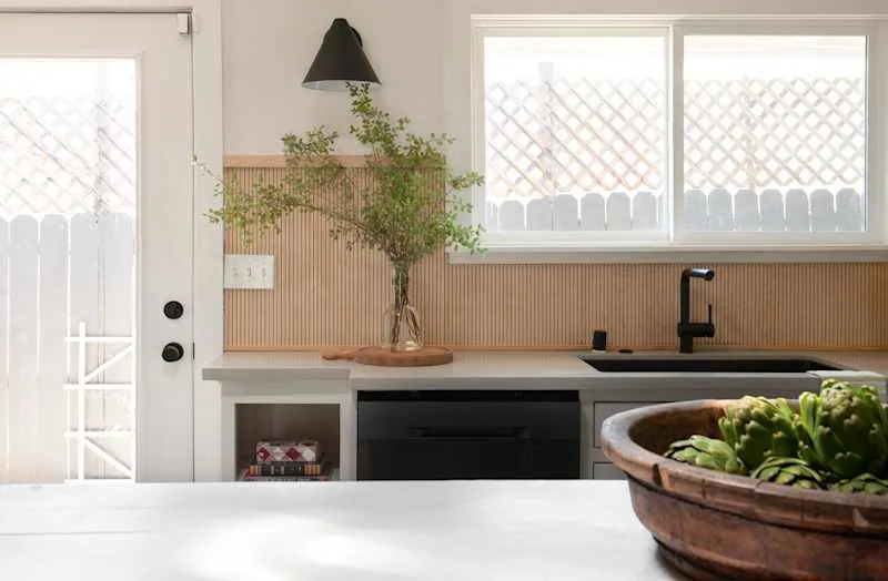 Kitchen with black faucet, wooden backsplash, potted plant and a bowl of artichokes on the countertop.