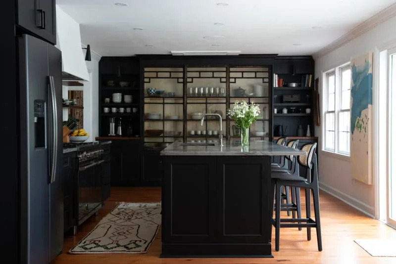 Modern kitchen with black cabinets, island with bar stools, glass-fronted shelves and a vase of white flowers.