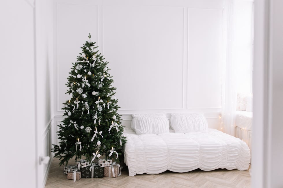 Cozy Christmas bedroom with white linen bed and decorated tree in Scandinavian style, bright comfortable room with Christmas decorations for winter season